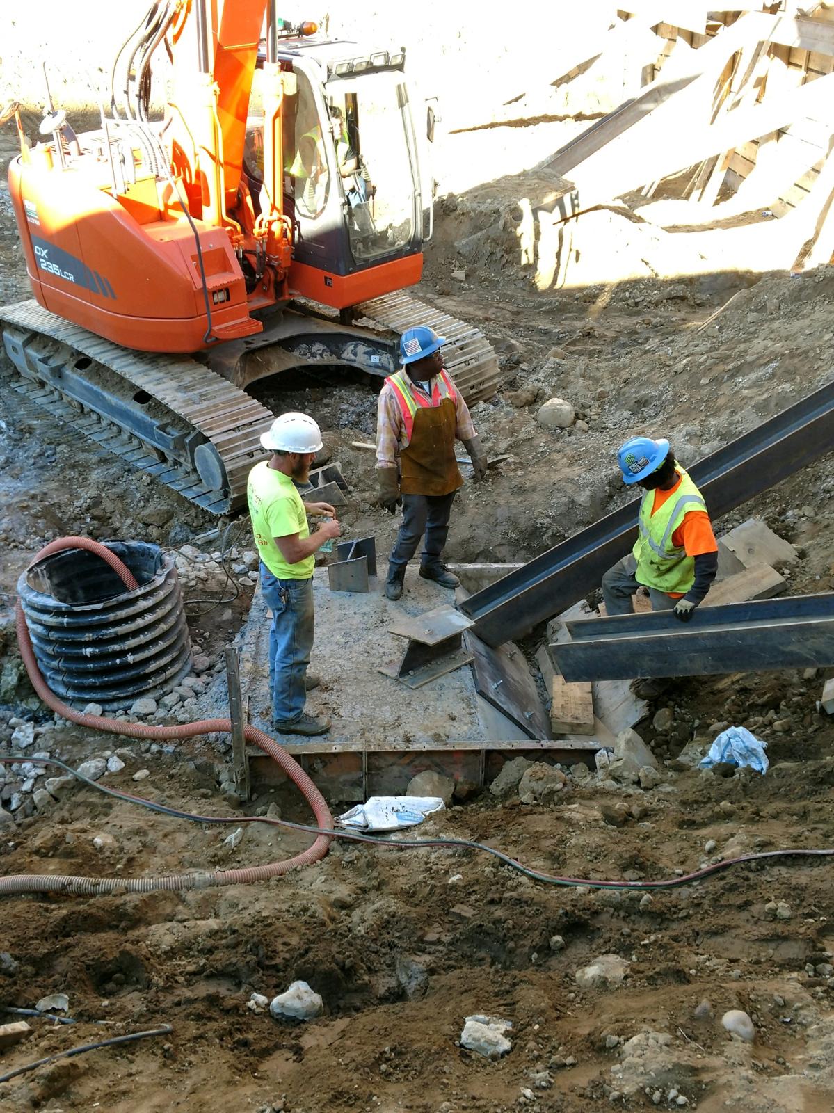 An ironworking crew setting a structural steel beam beside an excavator in an open excavation.
